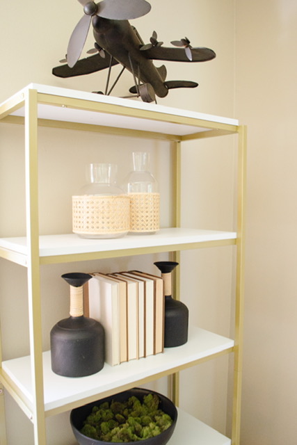 Gold and white bookshelf with iron plane model, cane vases, black vases with books and moss bowl. 